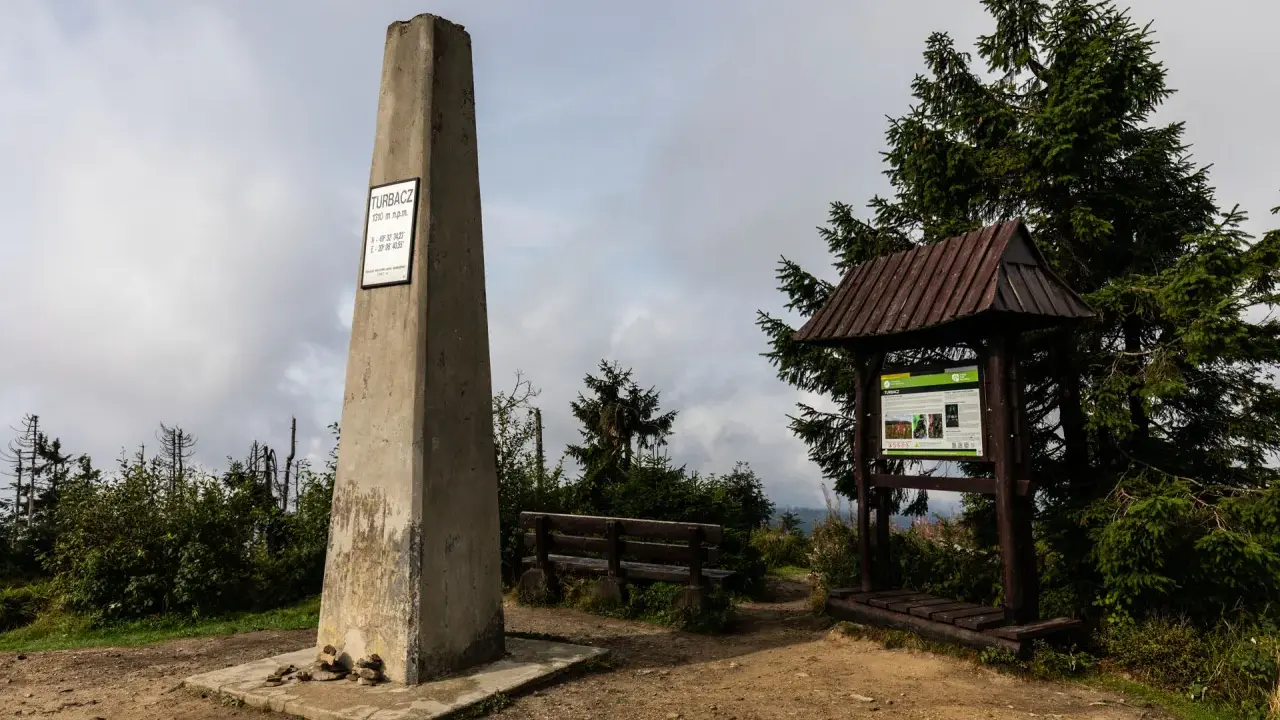 Kamienny obelisk na szczycie Turbacz w Gorcach, obok drewniana wiata z tablicą informacyjną i ławką.