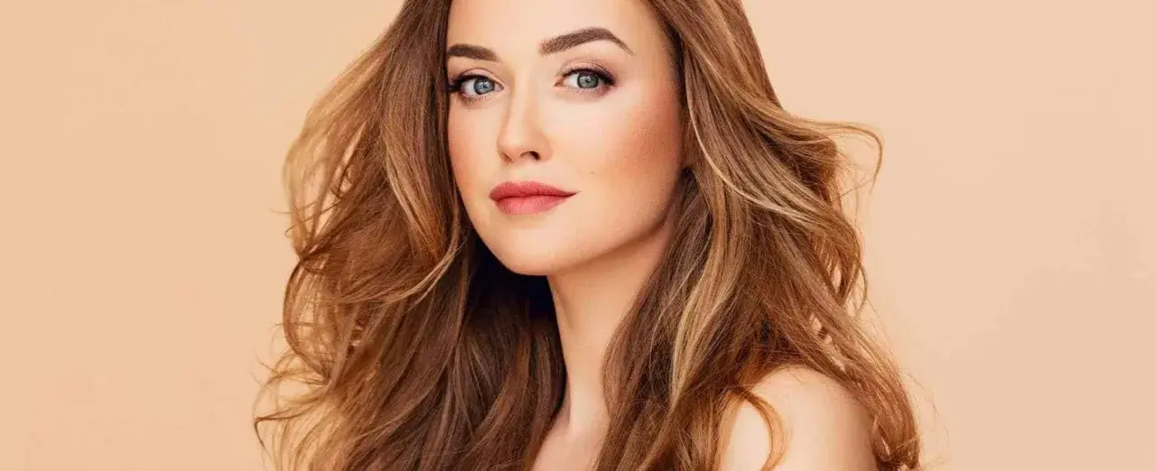 Mujer con cabello castaño dorado, ondulado y con reflejos claros. Su mirada es penetrante y sus labios pintados de rosa.