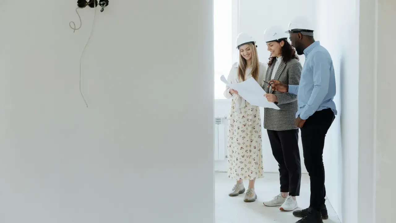 Tres arquitectos con cascos revisan planos en una obra, buscando cómo abaratar costes.