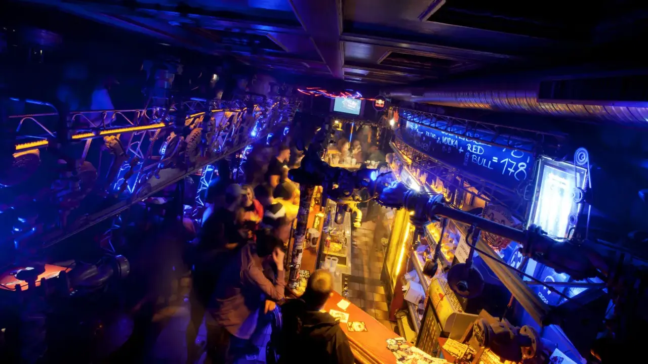 Lebhafte Nacht in einem der Prager Clubs, beleuchtet von blauem und gelbem Licht. Menschen stehen an der Bar und unterhalten sich.