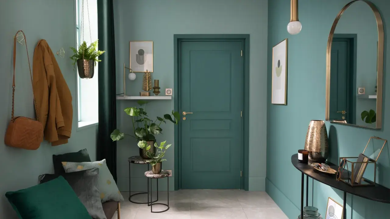 Entrada decorada con pared verde azulado, puerta verde, perchero con abrigo y bolso, plantas y espejo.