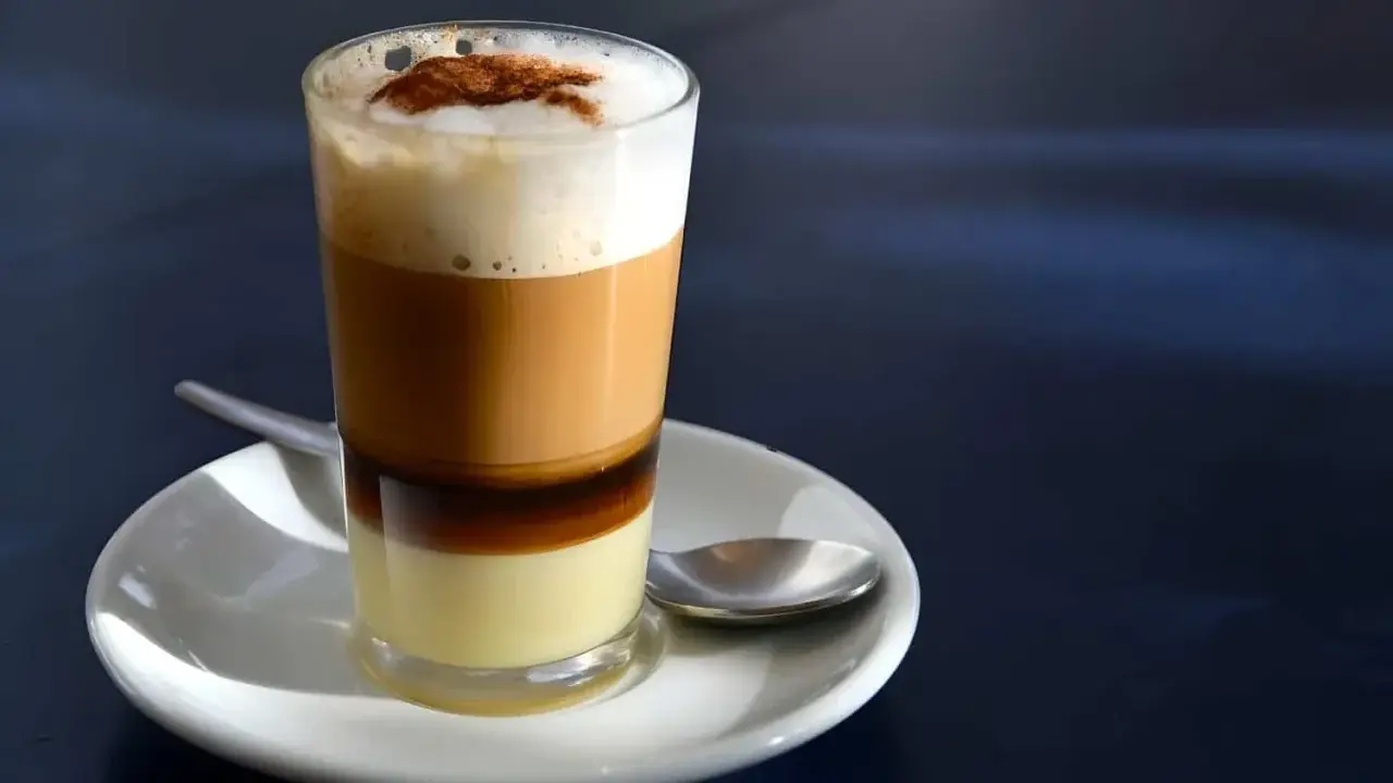 Ein geschichteter Kaffee con leche in einem Glas, mit einer Löffel auf einer Untertasse.