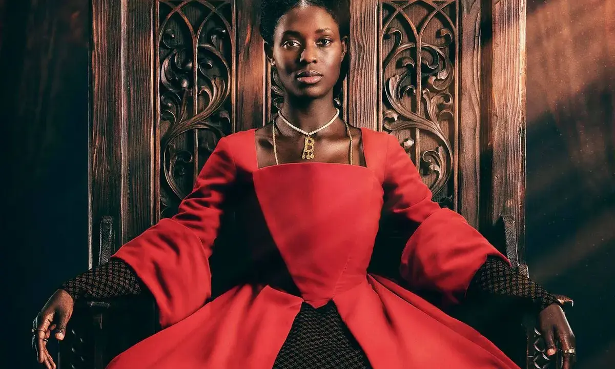 Jodie Turner-Smith, con su mirada regia, se sienta en un trono de madera tallada, vestida con un opulento traje rojo.