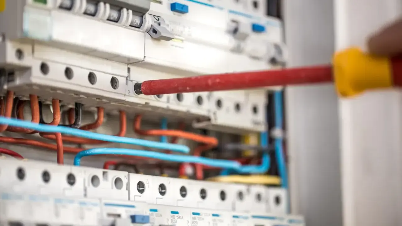 Ręka z czerwonym śrubokrętem pracuje przy instalacji niskoprądowej. Widać bezpieczniki i kolorowe przewody.