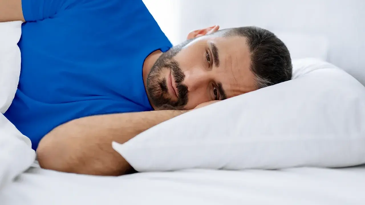 Hombre con barba en cama, con ojos abiertos, vistiendo camiseta azul. Duermo pero no descanso, ¿qué puedo hacer?