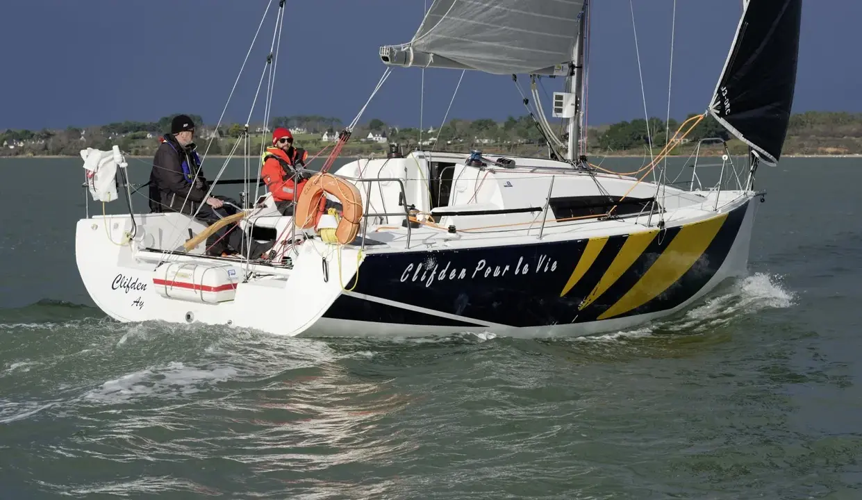Voile rapide, le voilier "Clifden Pour la Vie" navigue sur une mer agitée. Deux personnes sont à bord, profitant du soleil.
