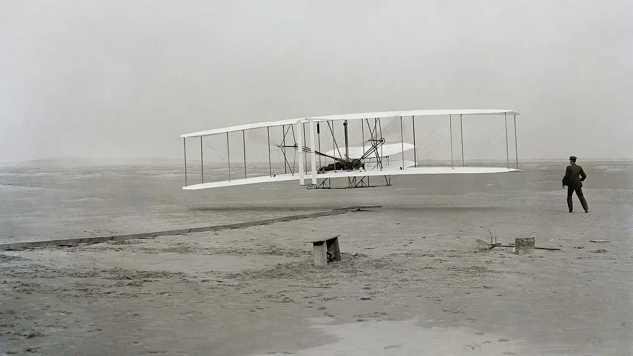 Der Wright Flyer hebt vom sandigen Boden ab, ein Mann beobachtet den historischen Flug.