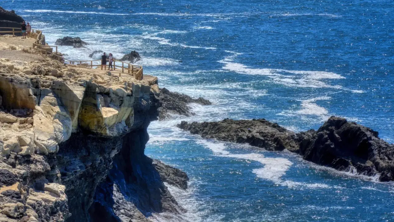 Acantilados rocosos y el mar azul cerca de las cuevas de Ajuy. Turistas observan el paisaje desde un mirador.