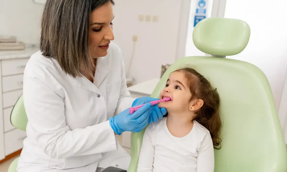 Dentystka w białym fartuchu i niebieskich rękawiczkach pokazuje małej dziewczynce, jak prawidłowo szczotkować zęby. To przykład troski o zdrowie jamy ustnej w ramach pedodoncji.