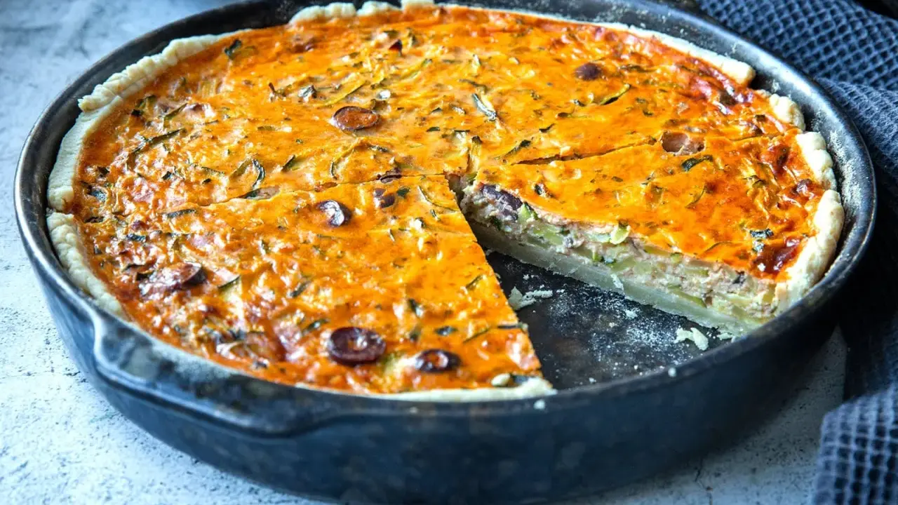Herzhaftes Quiche Rezept mit Zucchini und Oliven, goldbraun gebacken in einer Gusseisenpfanne. Ein Stück ist bereits herausgeschnitten.