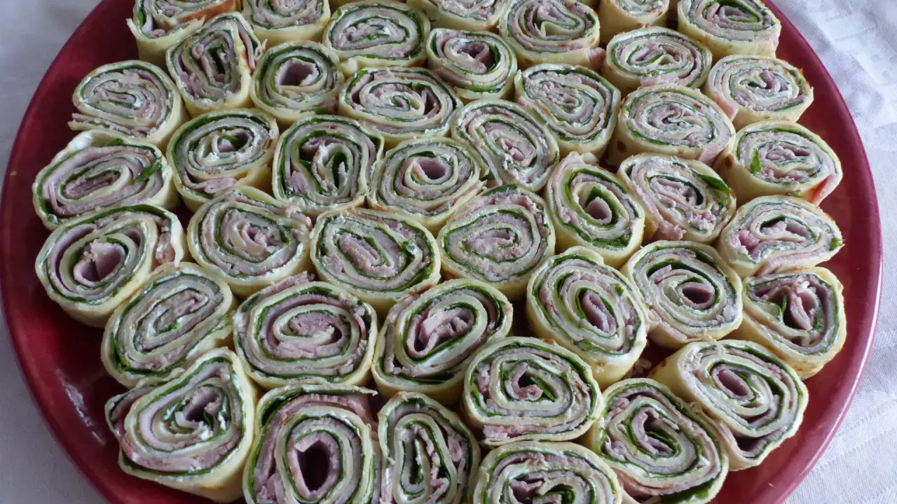 Une assiette rouge remplie de délicieuses crêpes roulées apéritif, garnies de jambon, fromage frais et herbes. Parfait pour un apéritif convivial.