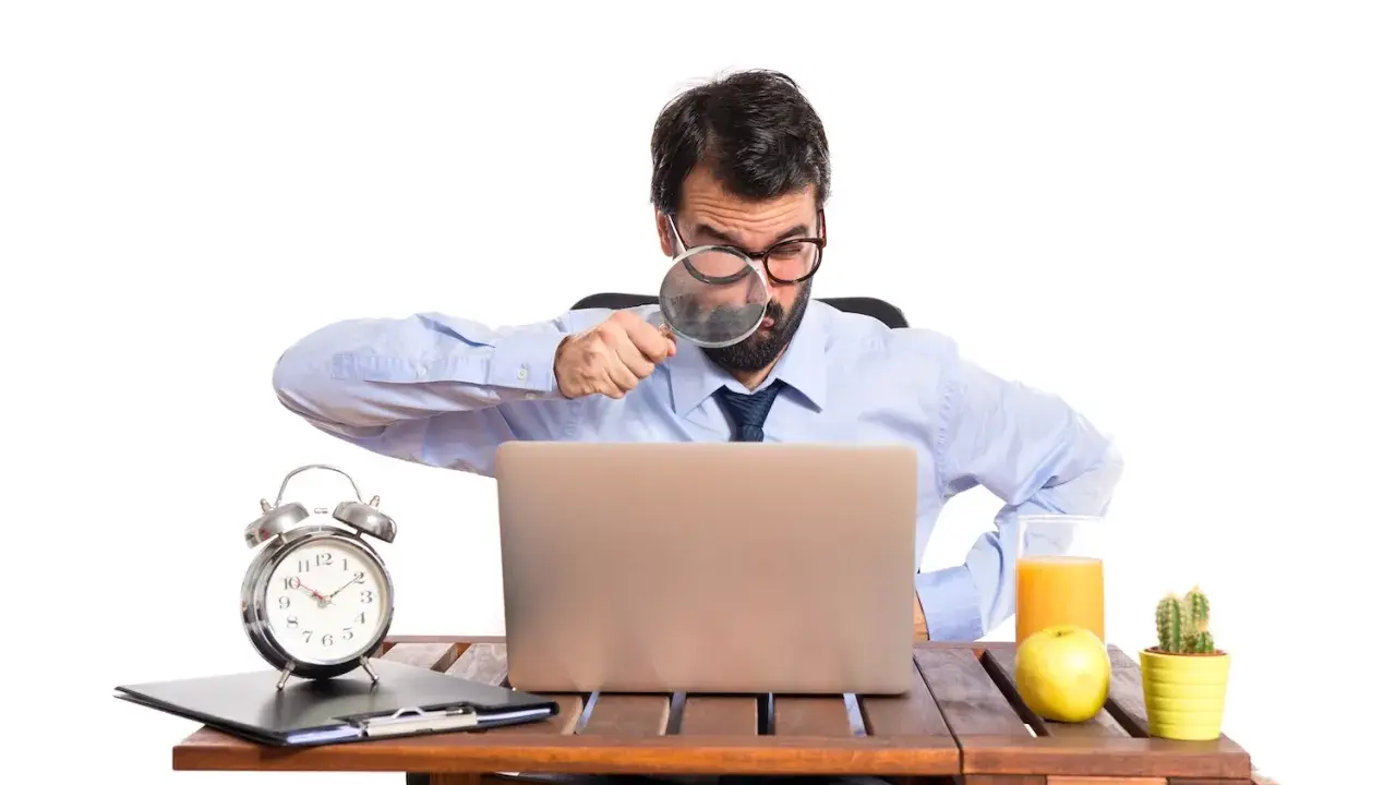 Mężczyzna z lupą bada laptopa, szukając odpowiedzi, czy skstore jest legit. Na biurku budzik, jabłko i sok.