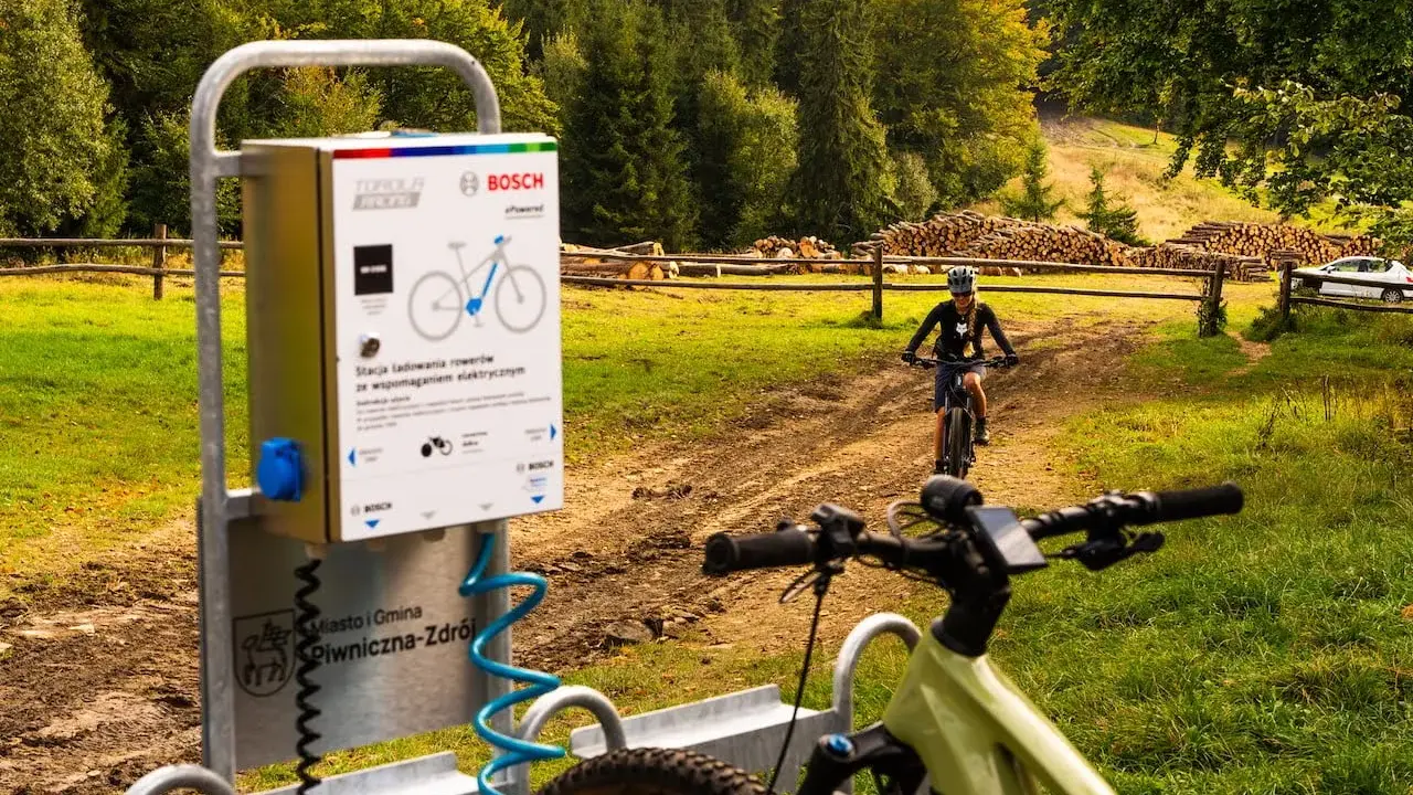 Stacje ładowania rowerów elektrycznych w Piwnicznej-Zdroju. W tle rowerzystka na szlaku.