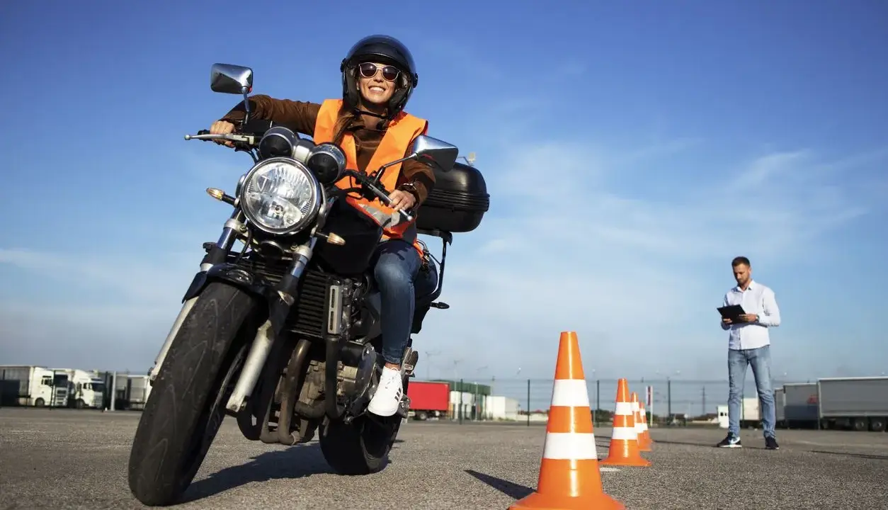 Mujer sonriente en moto, con chaleco naranja y casco, practicando en circuito con conos. Instructor observa. ¡Aprobando el examen de carbet a2!
