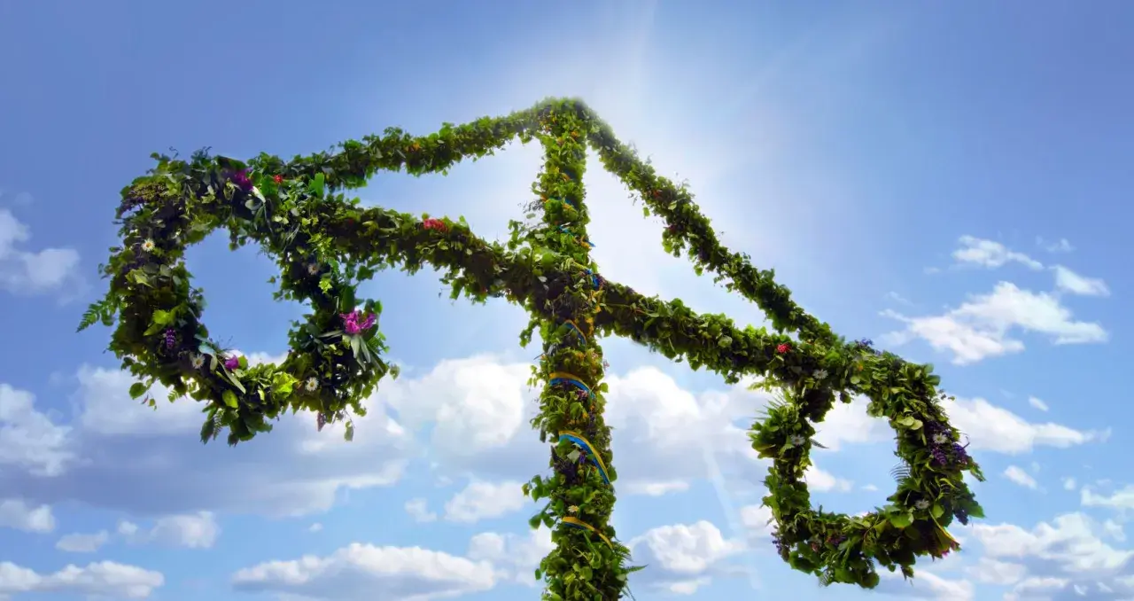 En midsommarstång, klädd i grönska och blommor, reser sig mot en klarblå himmel med solsken.