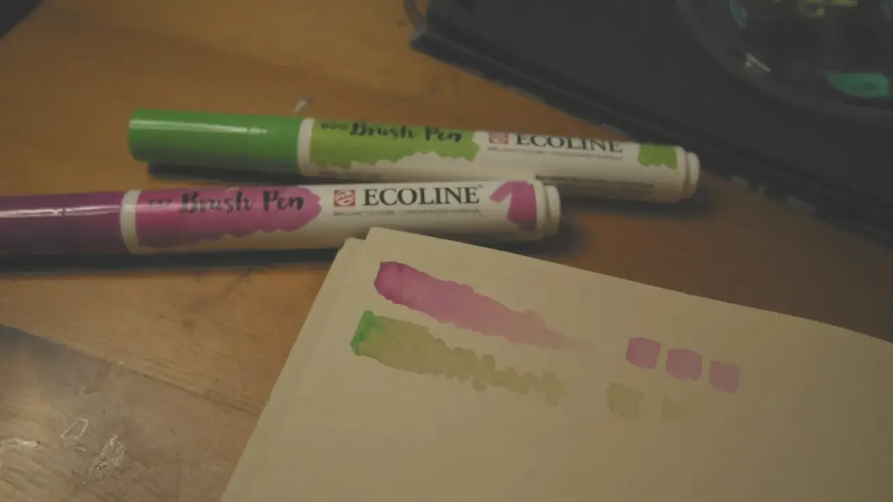 Dwa pisaki Ecoline (zielony i fioletowy) na drewnianym stole, obok próbki kolorów na papierze.