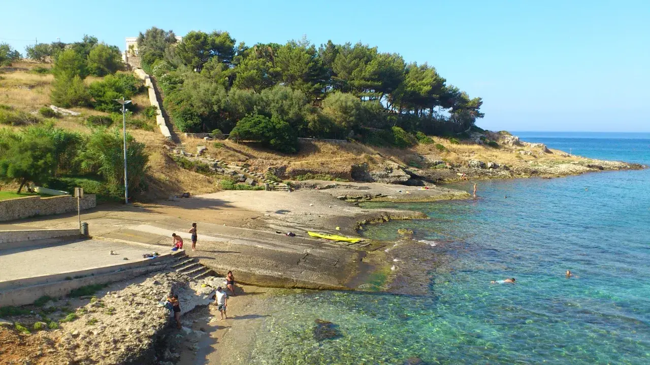 Scogliera e mare cristallino a San Gregorio Leuca. Persone si godono il sole e l'acqua, una scalinata sale verso la collina alberata.