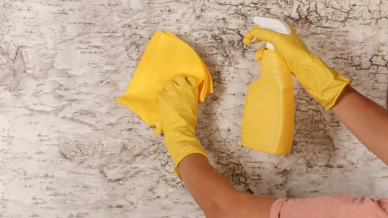 Manos con guantes amarillos limpiando paredes con un paño y spray.