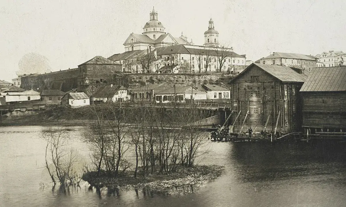 Widok na miasto z rzeką i kościołem. W oddali widać potężną budowlę sakralną z kopułami. Na pierwszym planie rzeka z wysepką i drzewami. Po prawej stronie drewniane budynki nad wodą. Ktoś pisze na Berdyczów.