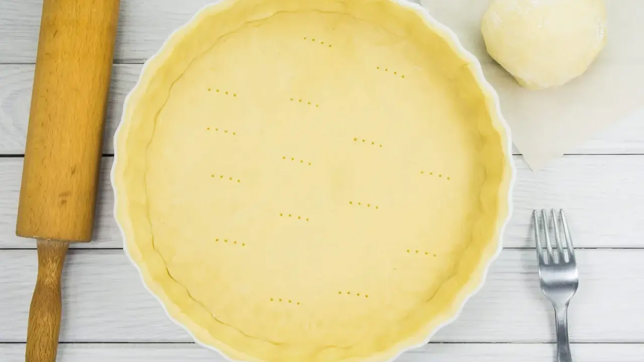 Une pâte brisée est prête dans une casserole blanche, avec une boule de pâte, une rouleau et une fourchette à côté.