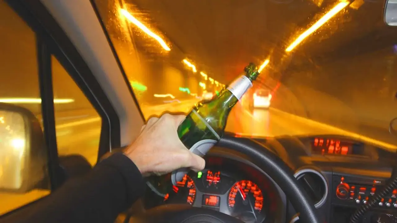 Ręka trzyma butelkę piwa za kierownicą. Po ilu można jechać autem po alkoholu? Nigdy!