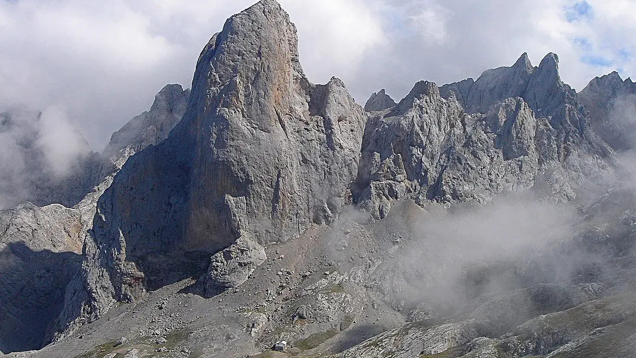 Majestatyczny **Naranjo de Bulnes** w Picos de Europa, otoczony mgłą i chmurami, z małym schroniskiem u podnóża.