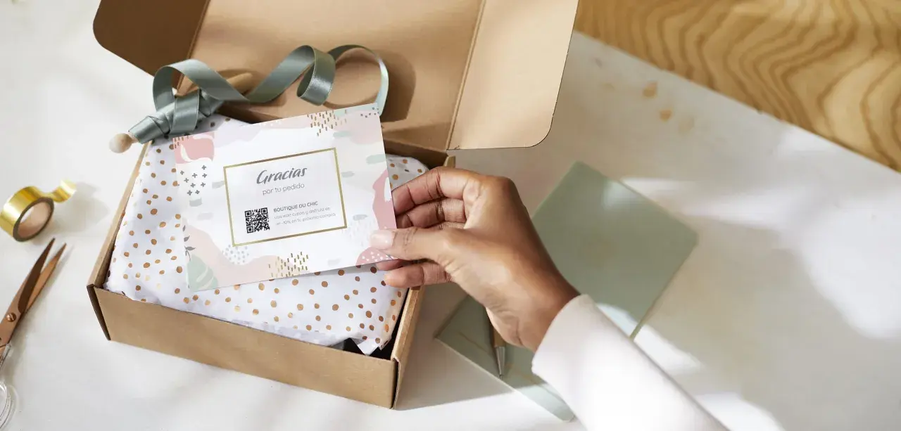 Tarjeta de agradecimiento tamaño postal con código QR, dentro de una caja de regalo abierta con papel de seda dorado.