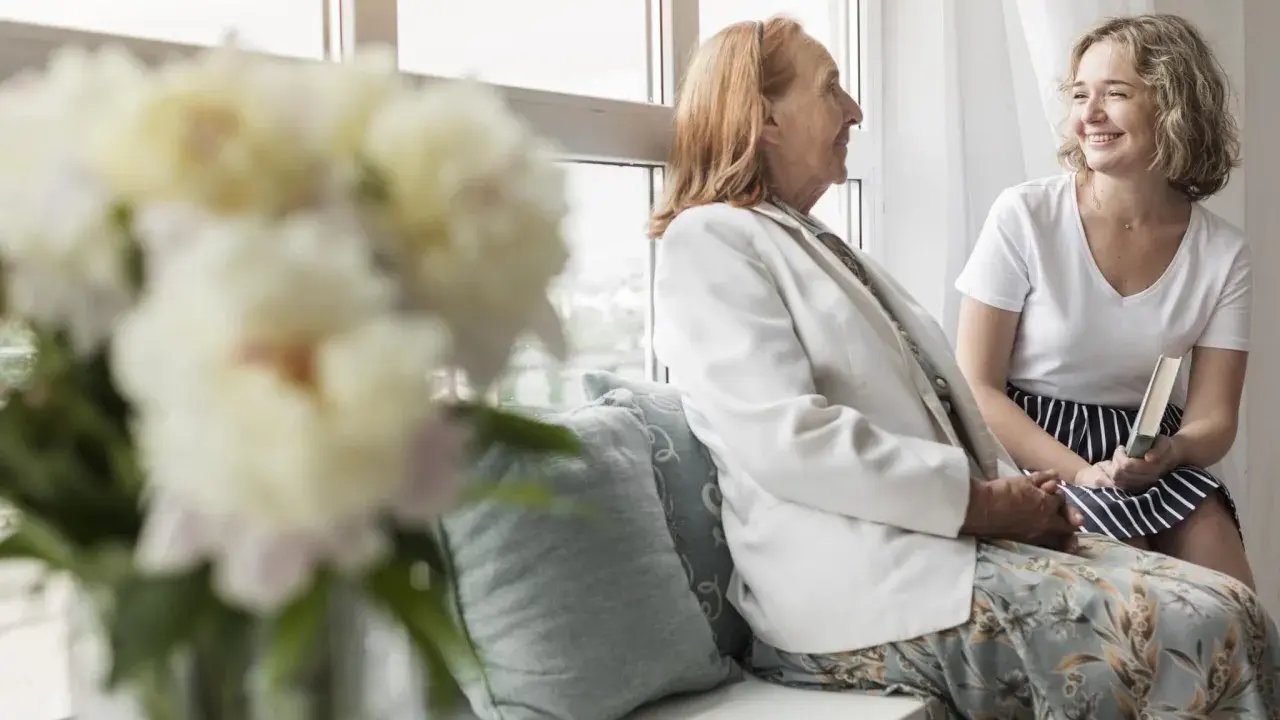 Uśmiechnięta młoda kobieta z książką rozmawia z seniorką przy oknie. Szukam pracy jako opiekunka prywatnie, oferując troskę i towarzystwo.