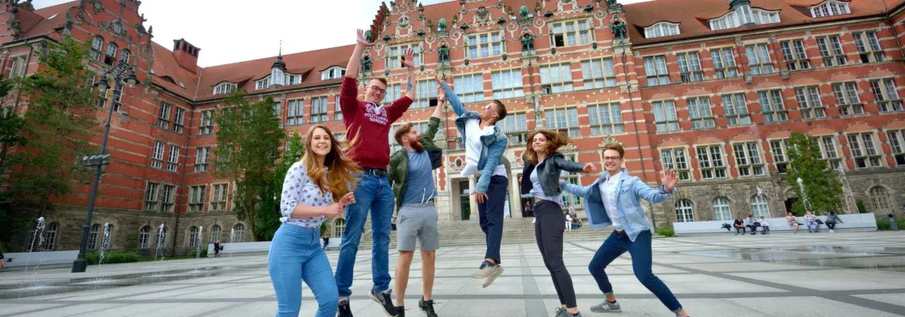 Radosna grupa studentów skacze przed budynkiem, gdzie można podjąć studia psychologiczne Gdańsk.