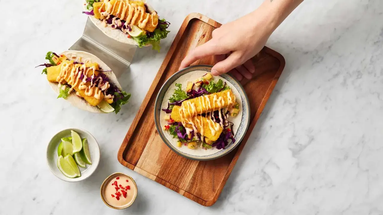 Två läckra fish tacos med krispig fisk, rödkål, sallad och en krämig sås. En hand sträcker sig efter en tallrik med en taco.
