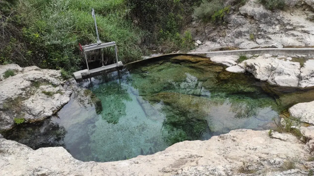 Aguas cristalinas en los baños de Somogil, rodeadas de rocas y vegetación. Una compuerta metálica controla el flujo del agua.