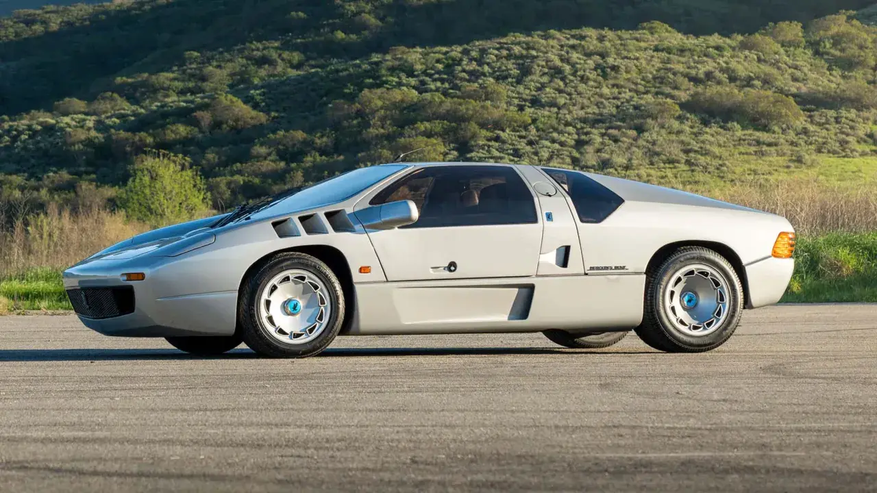 Silberner Isdera Imperator 108i mit markanten Lufteinlässen und futuristischem Design, geparkt vor einer grünen Hügellandschaft.
