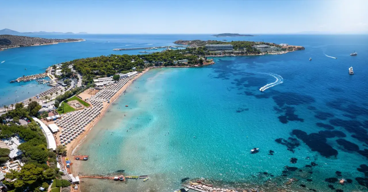 Traumhafter Blick auf das athen meer mit einer belebten Strandpromenade, Liegestühlen und dem glitzernden, türkisfarbenen Wasser, auf dem Boote fahren.