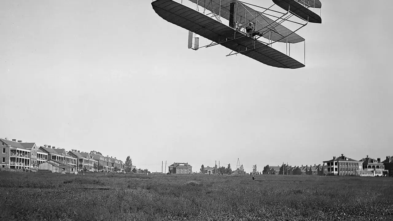 Das Flugzeug der Gebrüder Wright fliegt über ein Feld. Im Hintergrund sind Gebäude zu sehen.