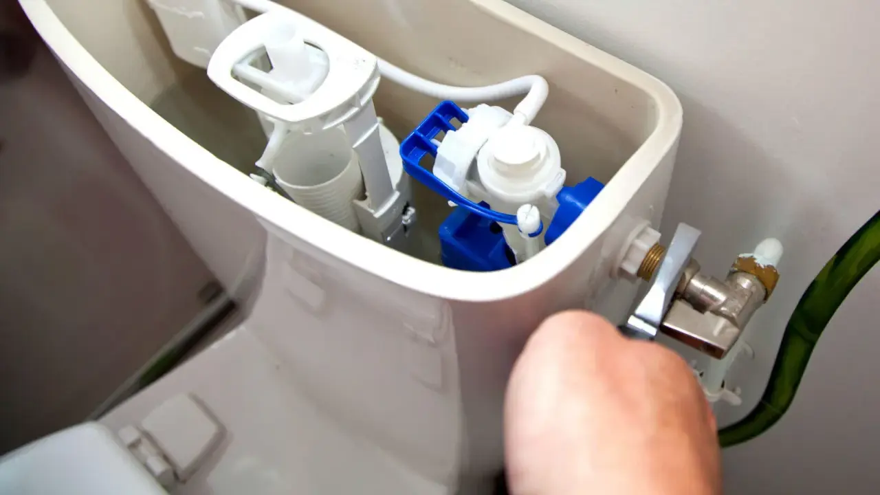 Mécanisme de chasse d'eau qui coule, avec un flotteur bleu et blanc visible. Une main ajuste le robinet d'arrivée d'eau.