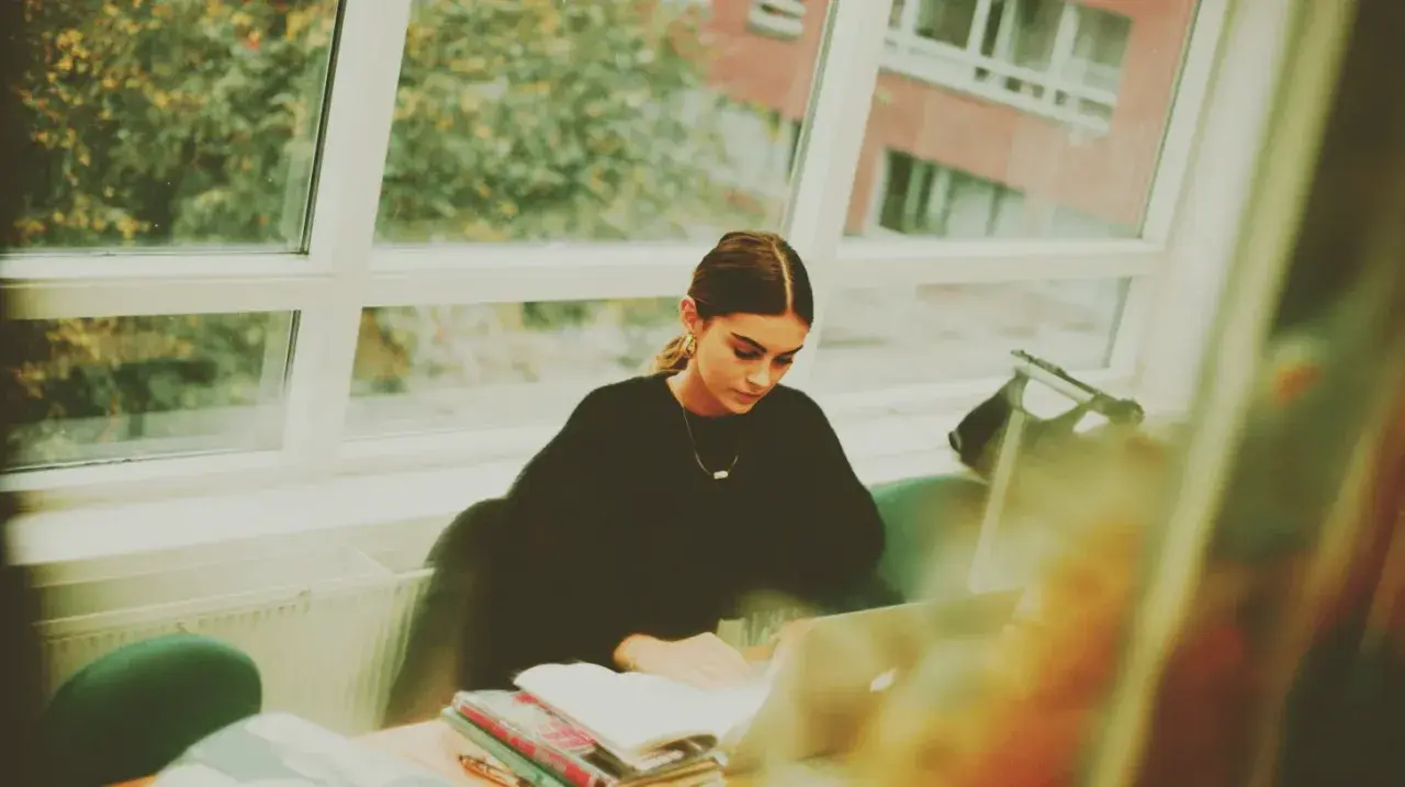 Studentka pochłonięta nauką kryminologii w Poznaniu, siedzi przy oknie z laptopem i książkami.