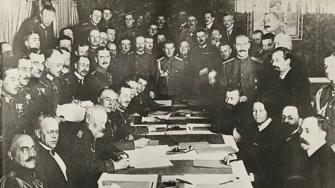 Delegierte unterzeichnen den Frieden von Brest-Litowsk. Männer in Uniformen und Anzügen sitzen an einem Tisch, umgeben von weiteren Personen.