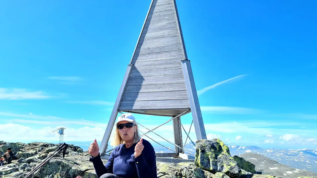 En kvinna sitter på toppen efter att ha lyckats bestiga Helags. Hon ger tummen upp vid den ikoniska träpyramiden.