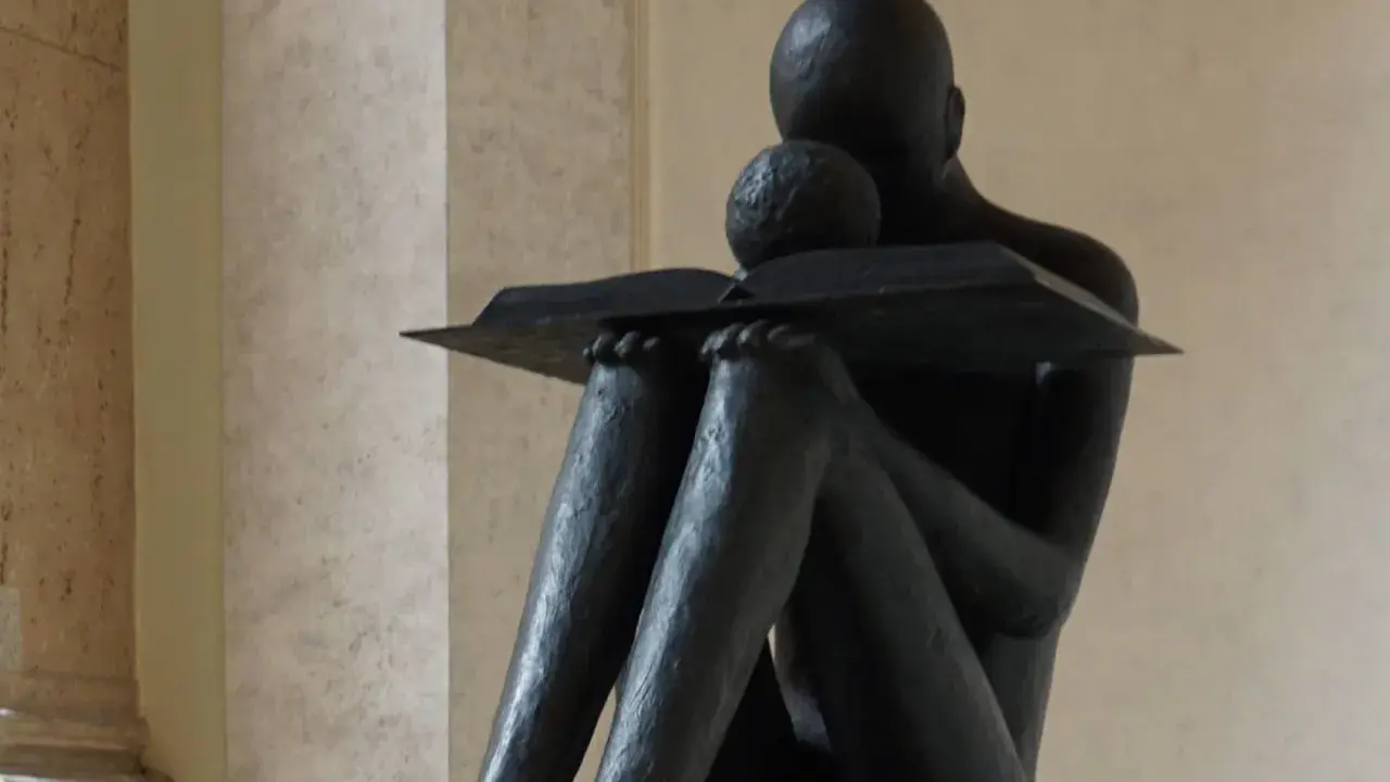 Statua di una figura che legge un libro, con un bambino appoggiato. Un'immagine che evoca il "sursum corda traduzione" della conoscenza.