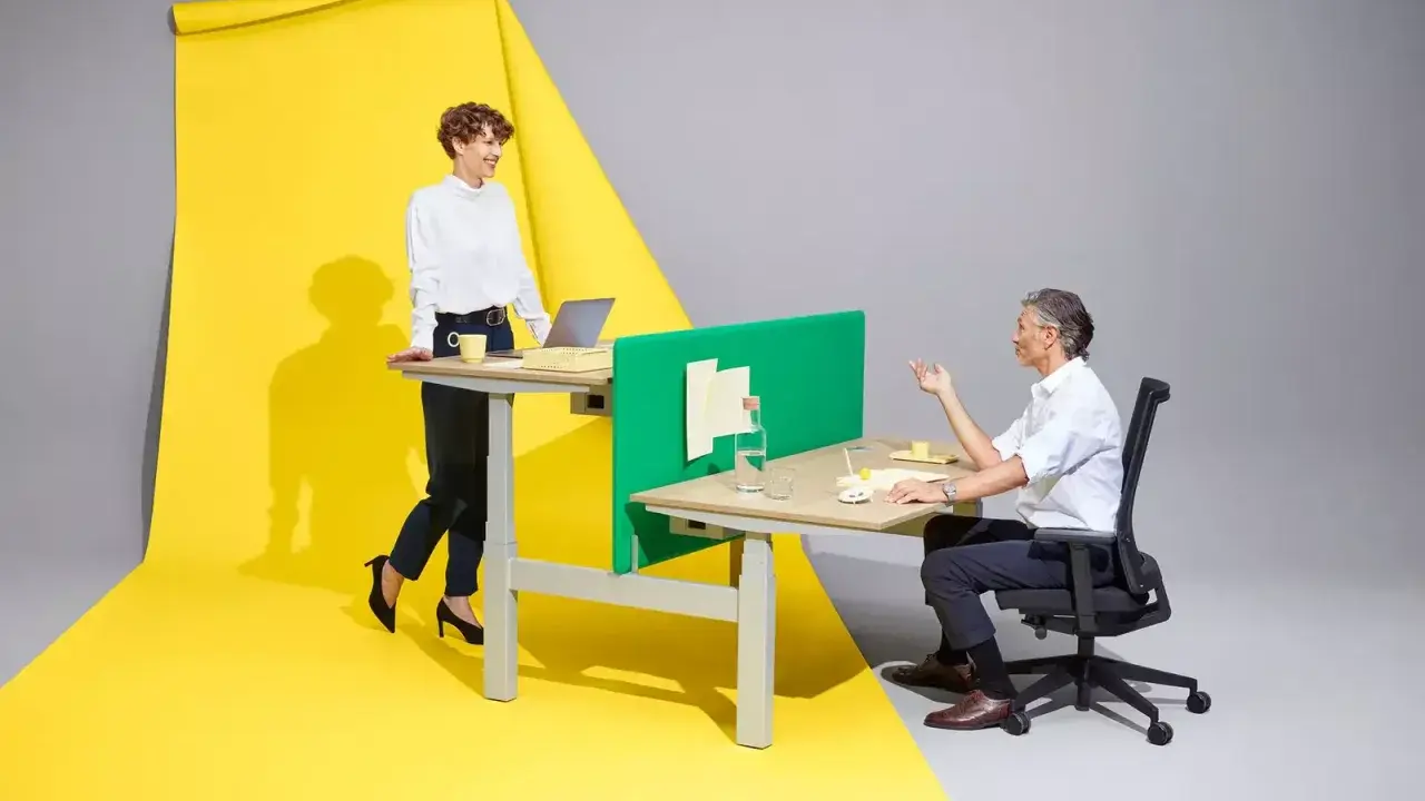 Femme en position debout statique, souriante, devant un bureau. Homme assis à un bureau, discutant. Fond jaune et gris.