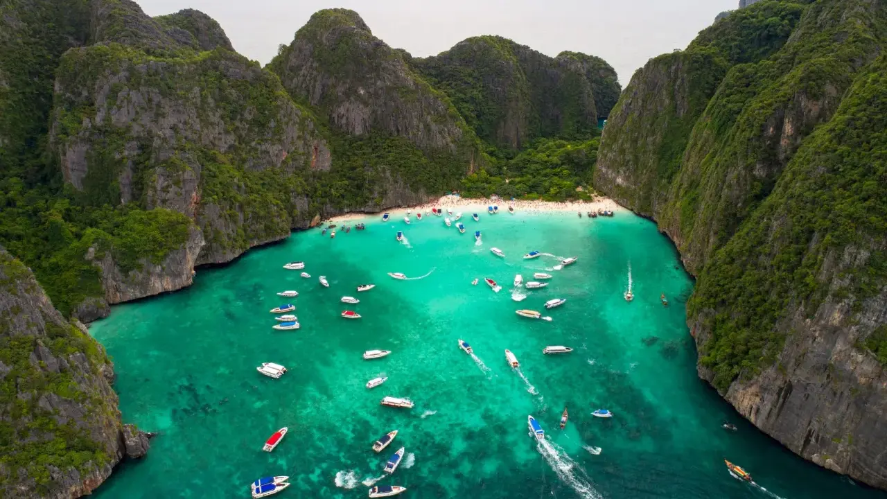 Maya Bay tętni życiem, pełna łodzi i turystów na piaszczystej plaży, otoczona stromymi, porośniętymi zielenią klifami.