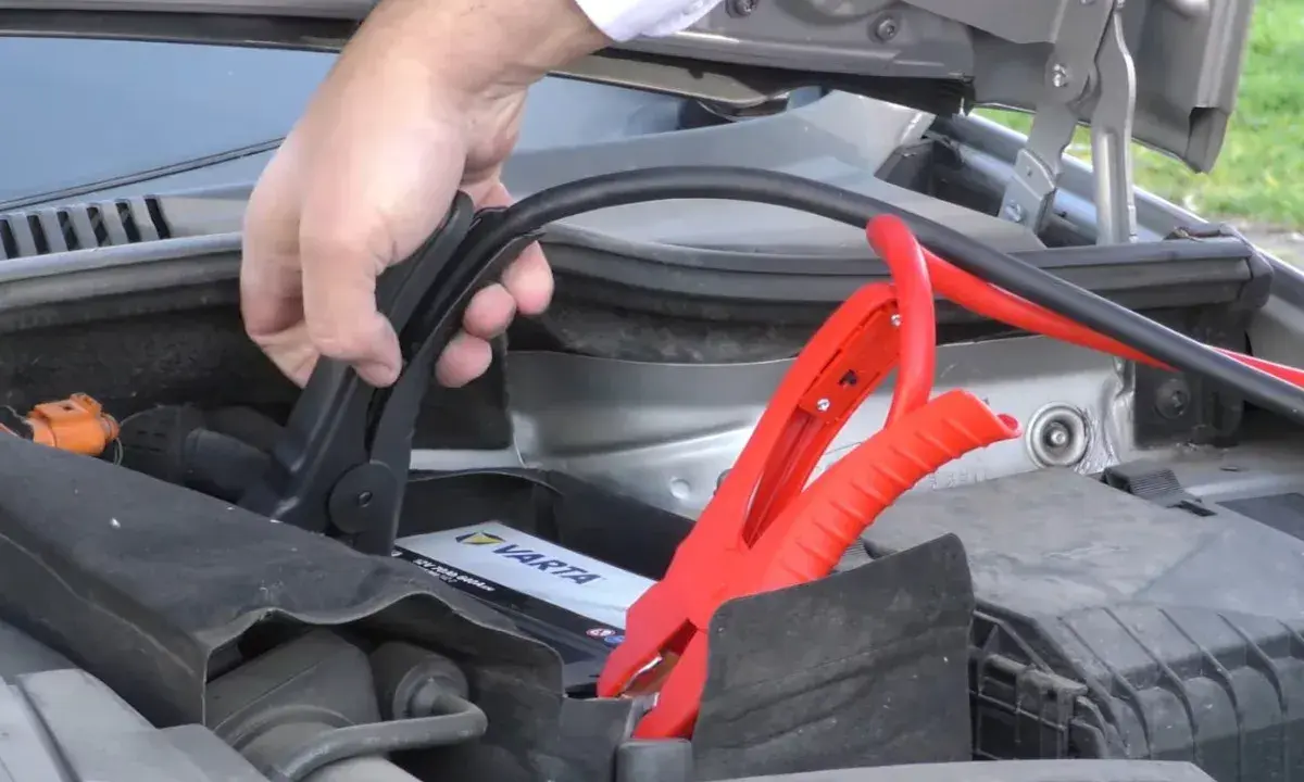 Mano conectando pinzas rojas a la batería de un coche para arrancar con pinzas.
