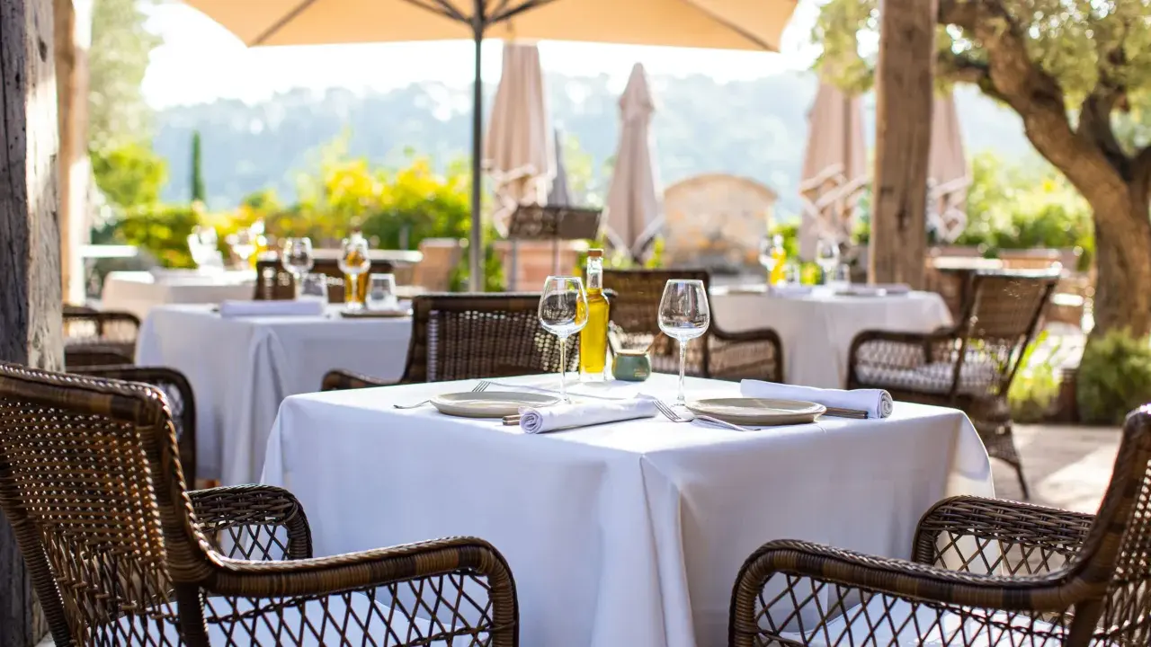 Tables dressées en terrasse, sous un parasol, invitant à savourer une cuisine du terroir authentique. Vins, verres et couverts attendent les convives.