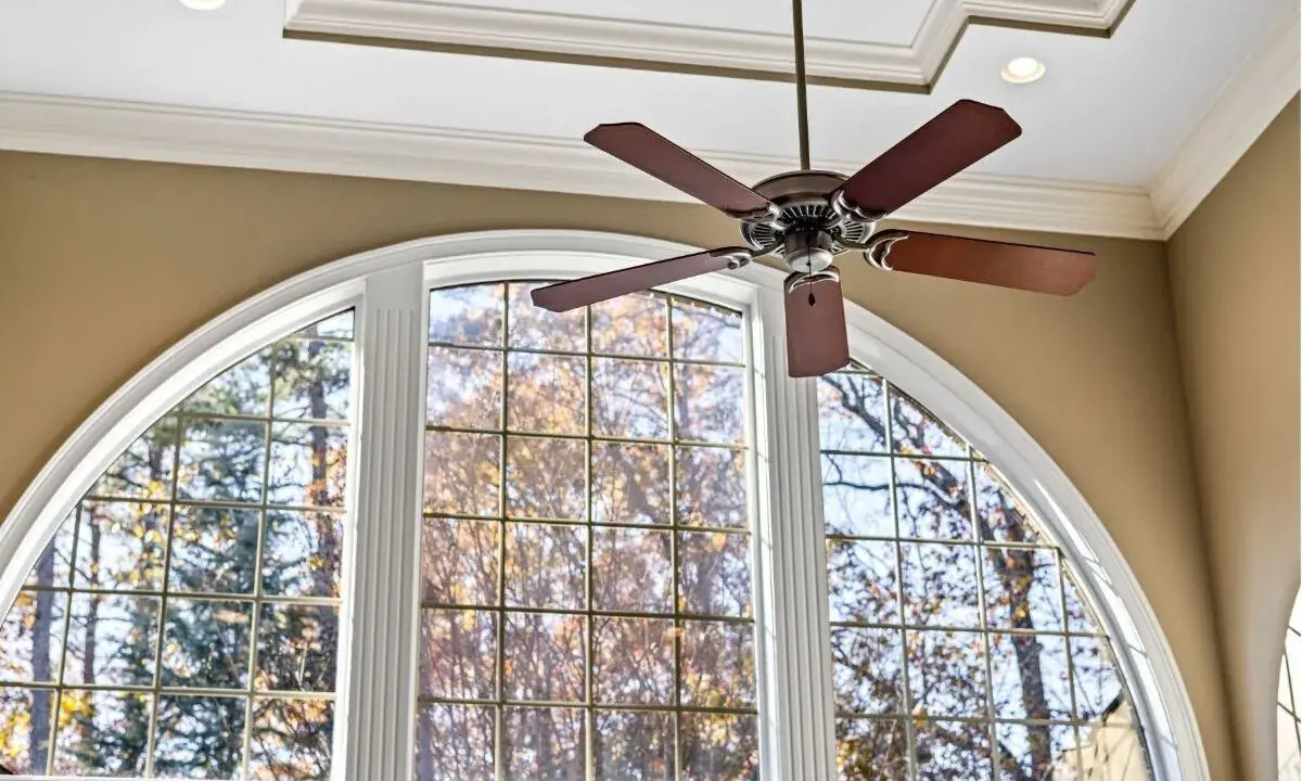Ventilador de techo de madera oscura sobre una ventana arqueada con vista a árboles otoñales.