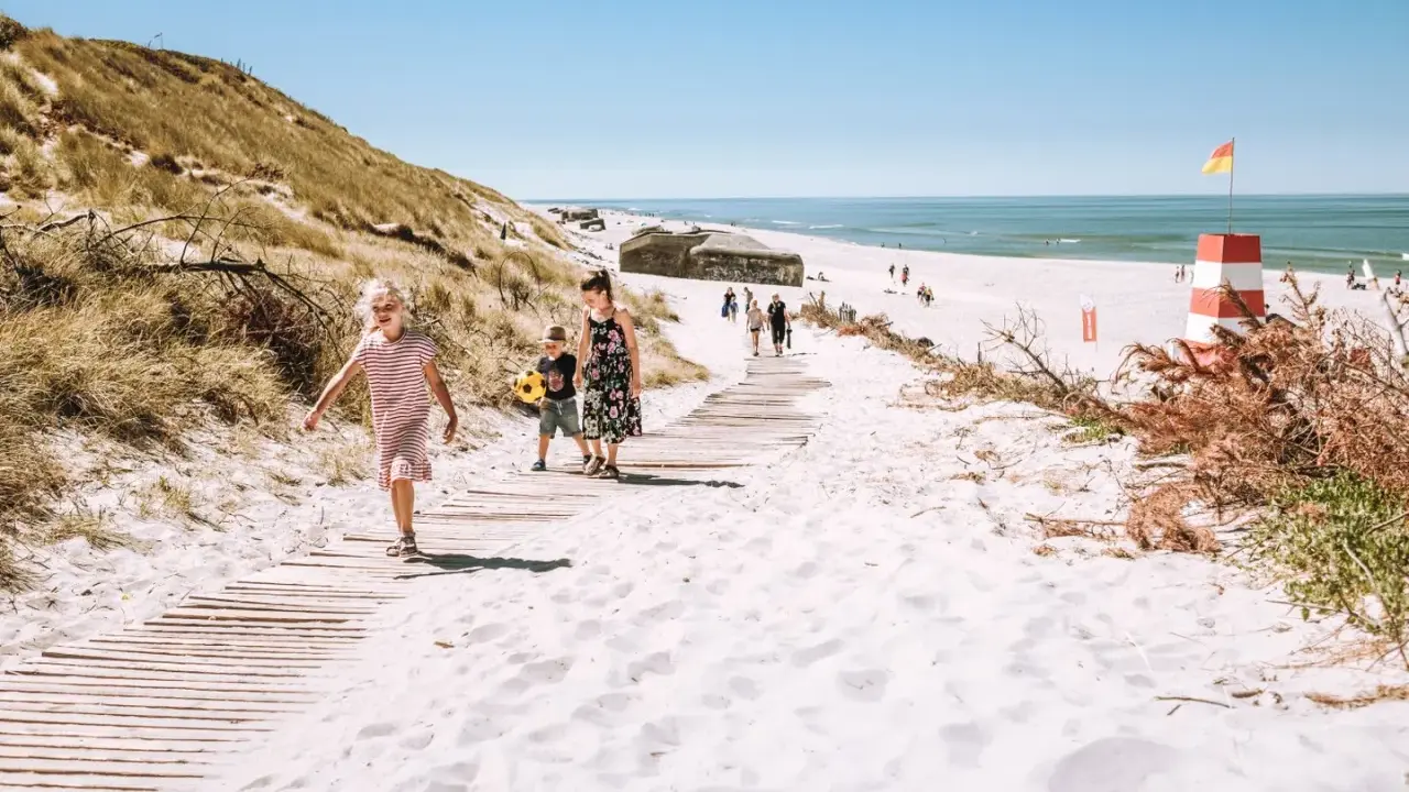 Familj på väg längs en träspång över vit sand i Danmark. En rödvit fyr och bunkrar syns i bakgrunden.