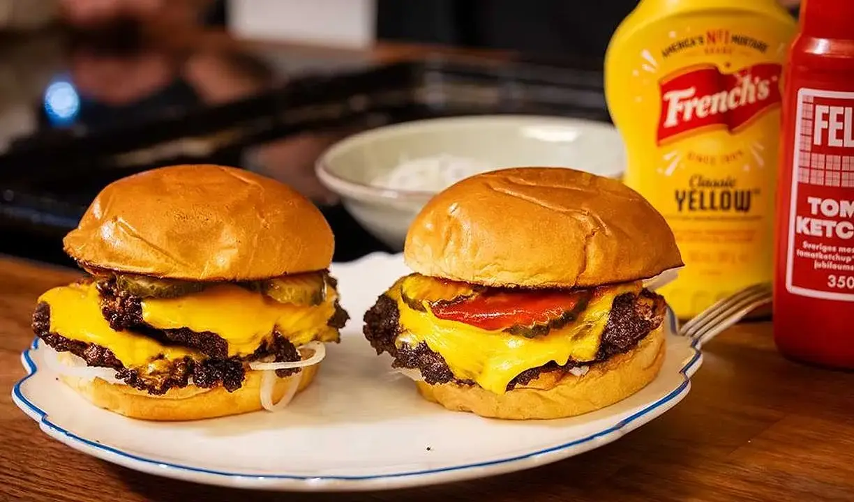 Två saftiga hamburgare med smält ost, pickles och lök på en tallrik. Bredvid står flaskor med senap och ketchup.