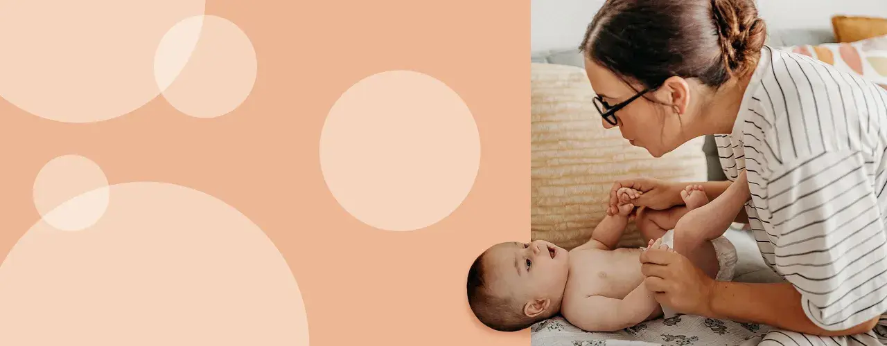 Mama z okularami delikatnie podnosi nóżki dziecka, przygotowując się do zakładania pampersa na stojąco.