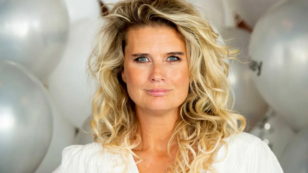 Blond kvinna med blå ögon och lockigt hår, omgiven av silverfärgade ballonger. Hon ser ut att vara redo för festliga udda veckor.