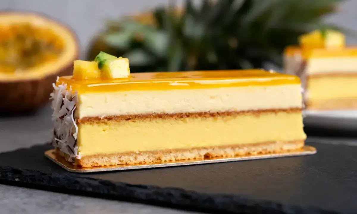 Un délice tropical : un entremets aux couches crémeuses, surmonté d'un glacage miroir jaune éclatant, décoré de fruits et de noix de coco râpées.