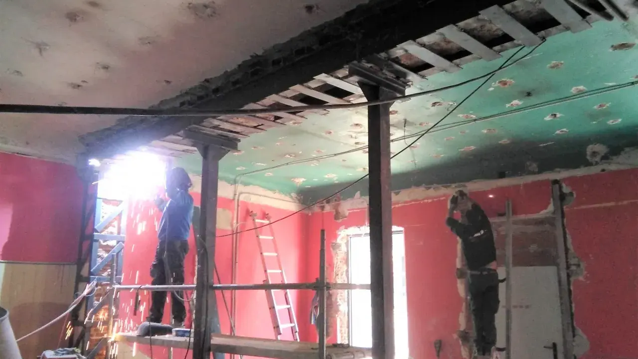 Trabajadores realizan el apeo de muro de carga con soportes metálicos y soldadura. El techo está en proceso de demolición.